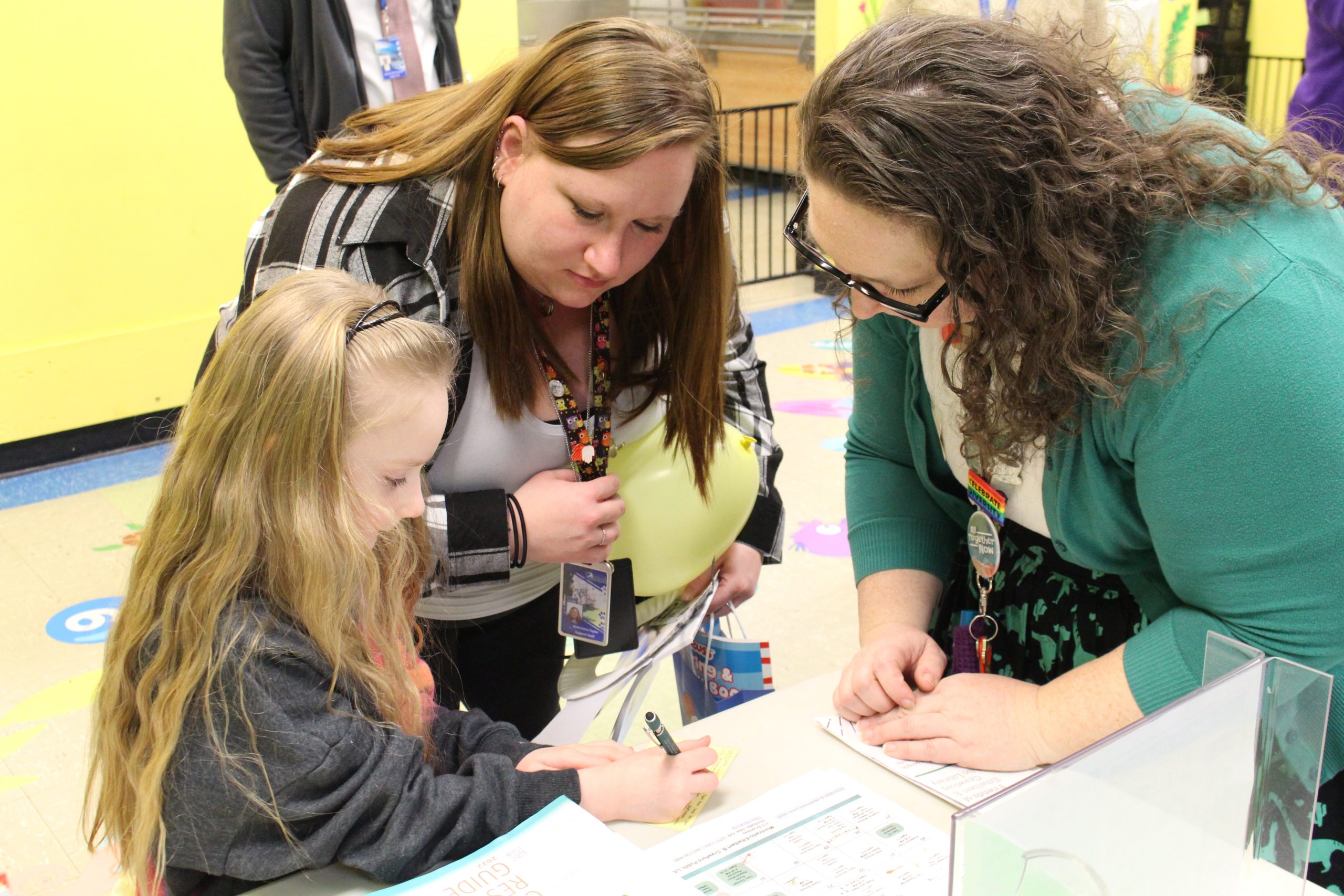 two adult women are huddled over a young girl as she fills out a library card application 