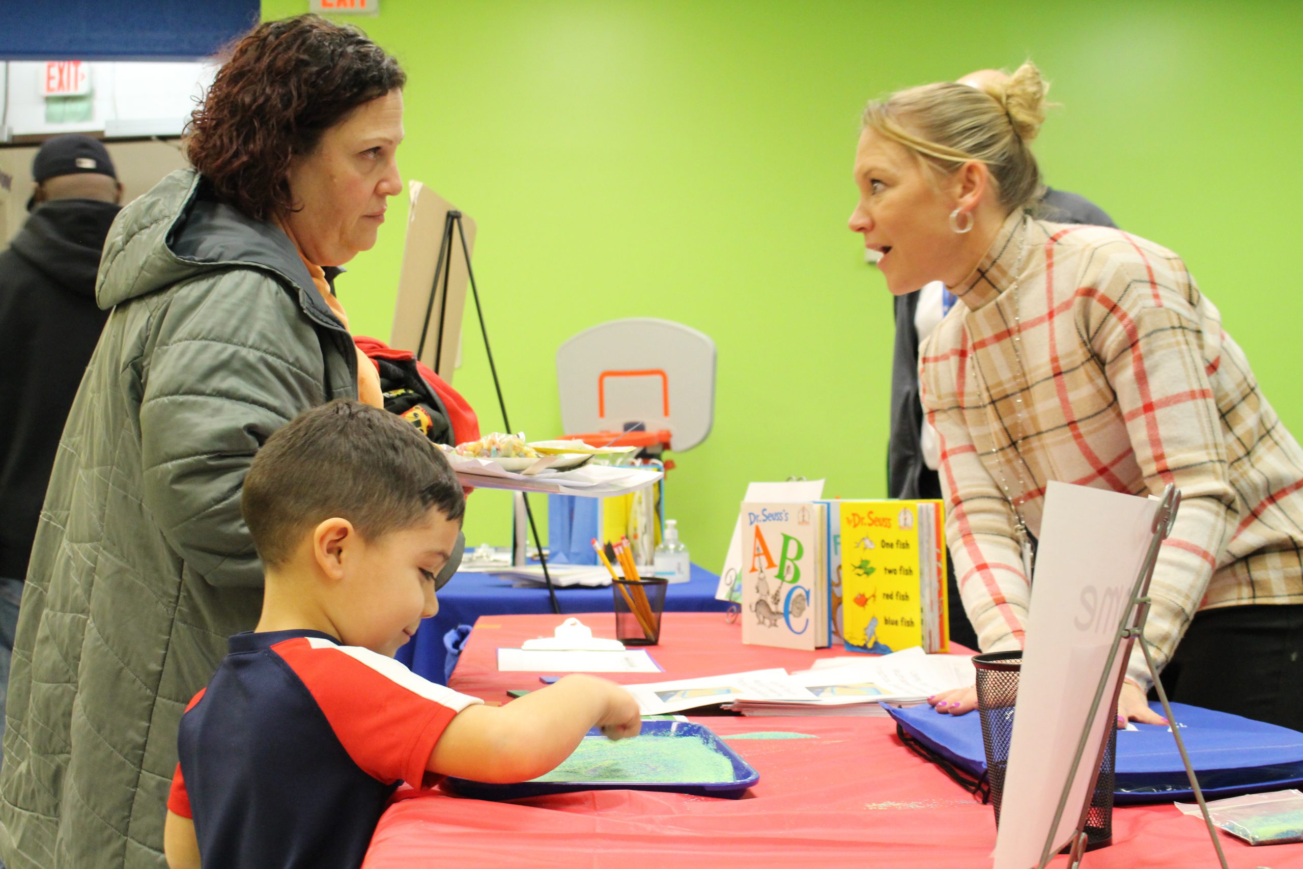 two adult women are speaking across a table while a young boy traces shapes in sand. 