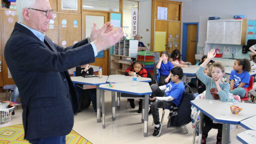 Mr. Conway is standing before a classroom of students. He has his hands out as if gesturing. The students are all seated at desks listening intently.