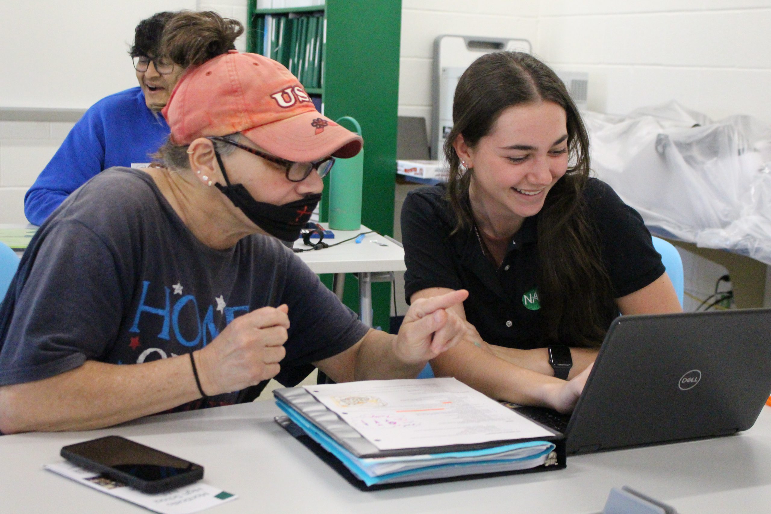 a student is helping a female community member. They are seated at a desk and the student is pointing at a computer. 