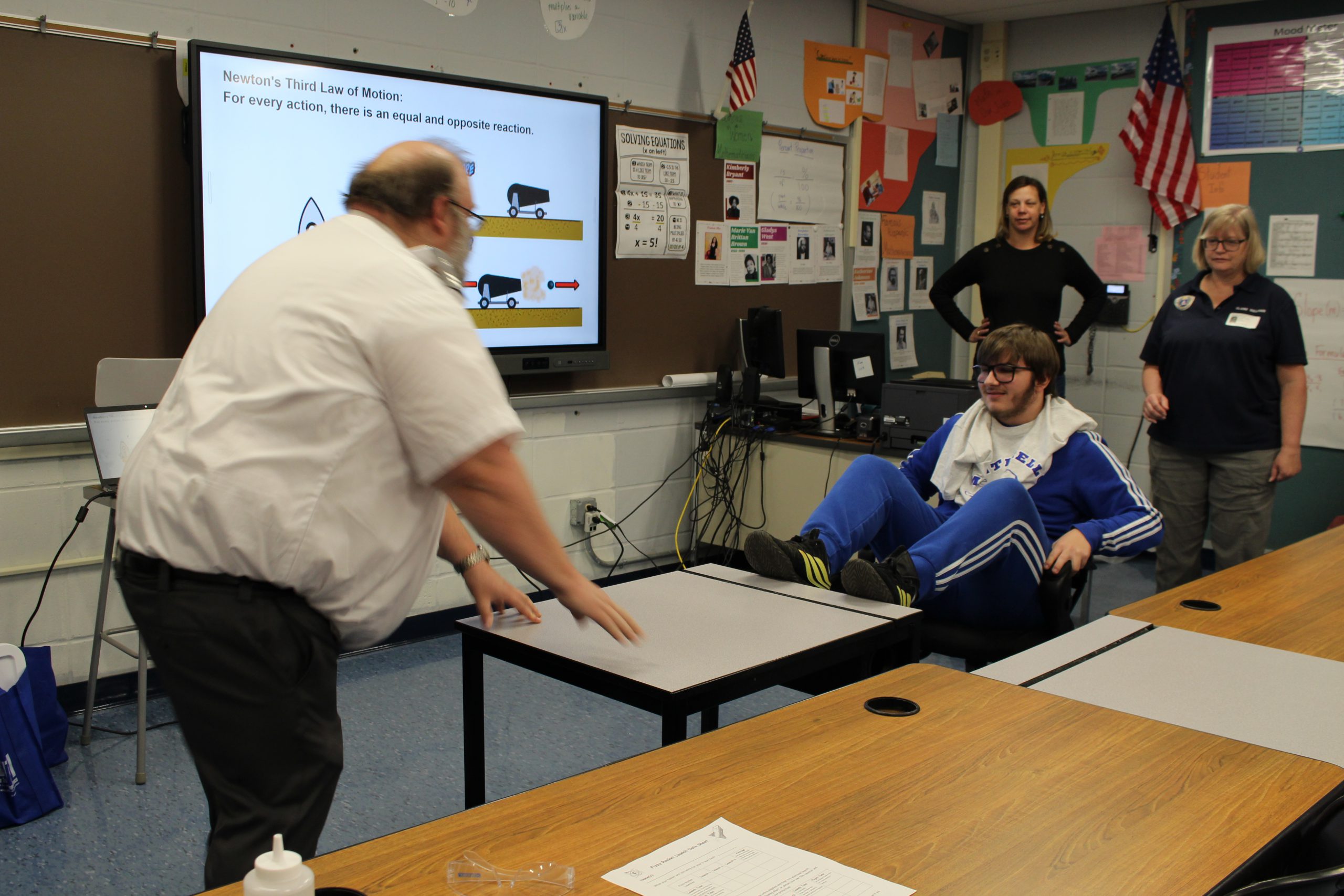 a student is seated in a chair and is using his legs to push a desk towards a man. The man is pushing the desk back towards the student. It's part of a experiment designed to demonstrate the third law of motion