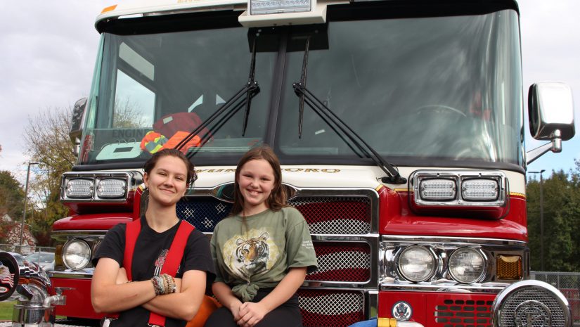 A female firefighter is learning against a firetruck next to a student who is seated on the grill.