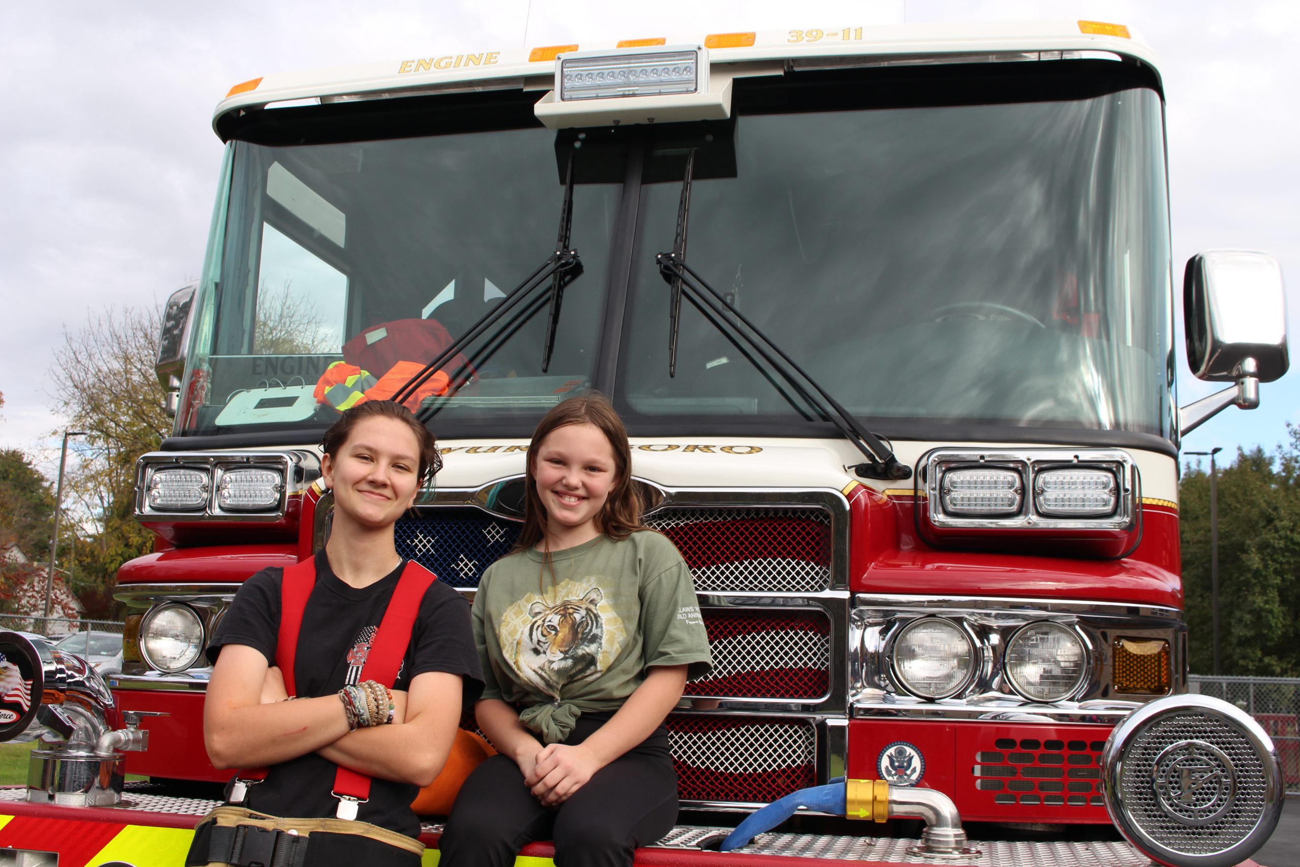 A female firefighter is learning against a firetruck next to a student who is seated on the grill. 
