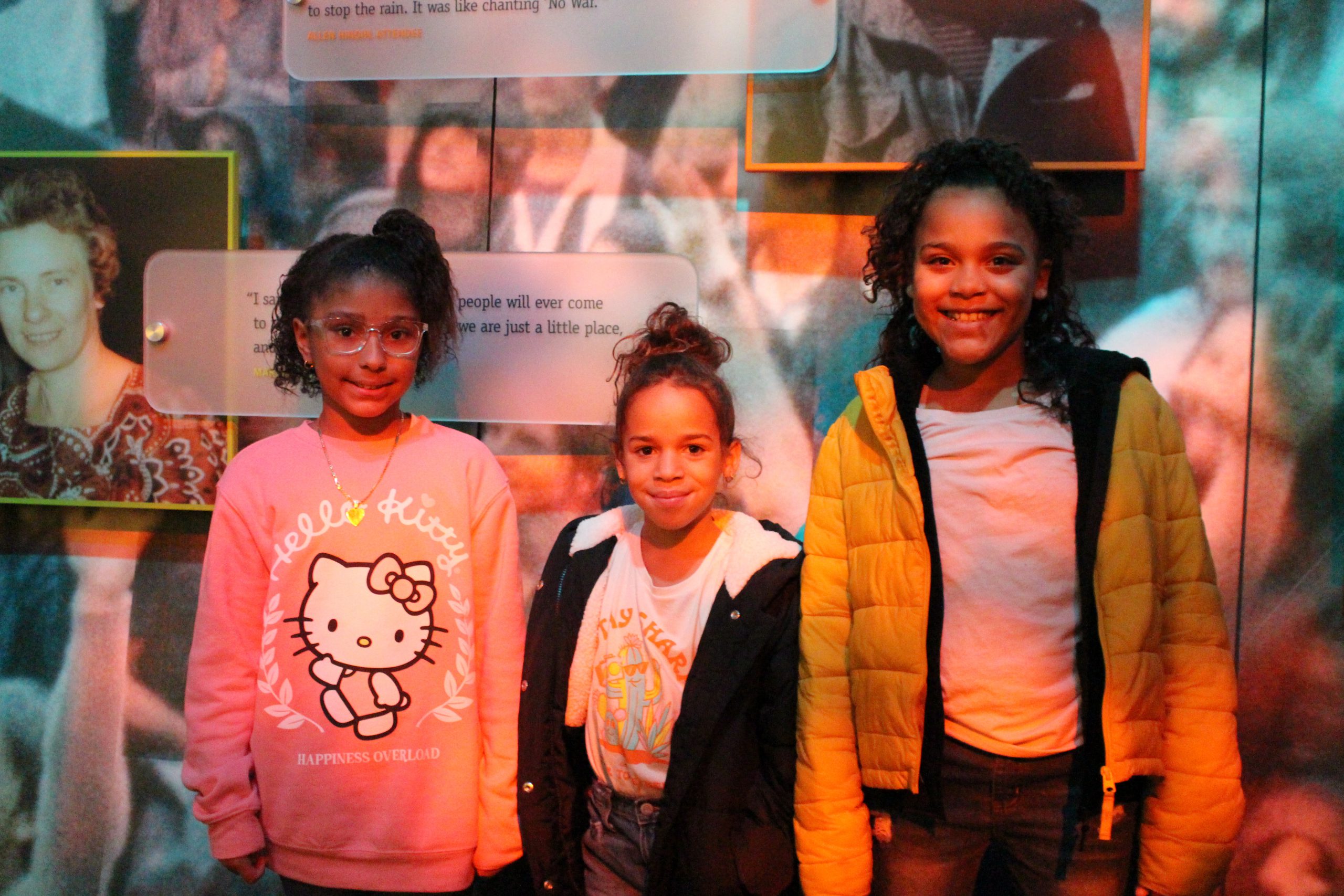 three girls are standing in front of Woodstock memorabilia at the museum. 