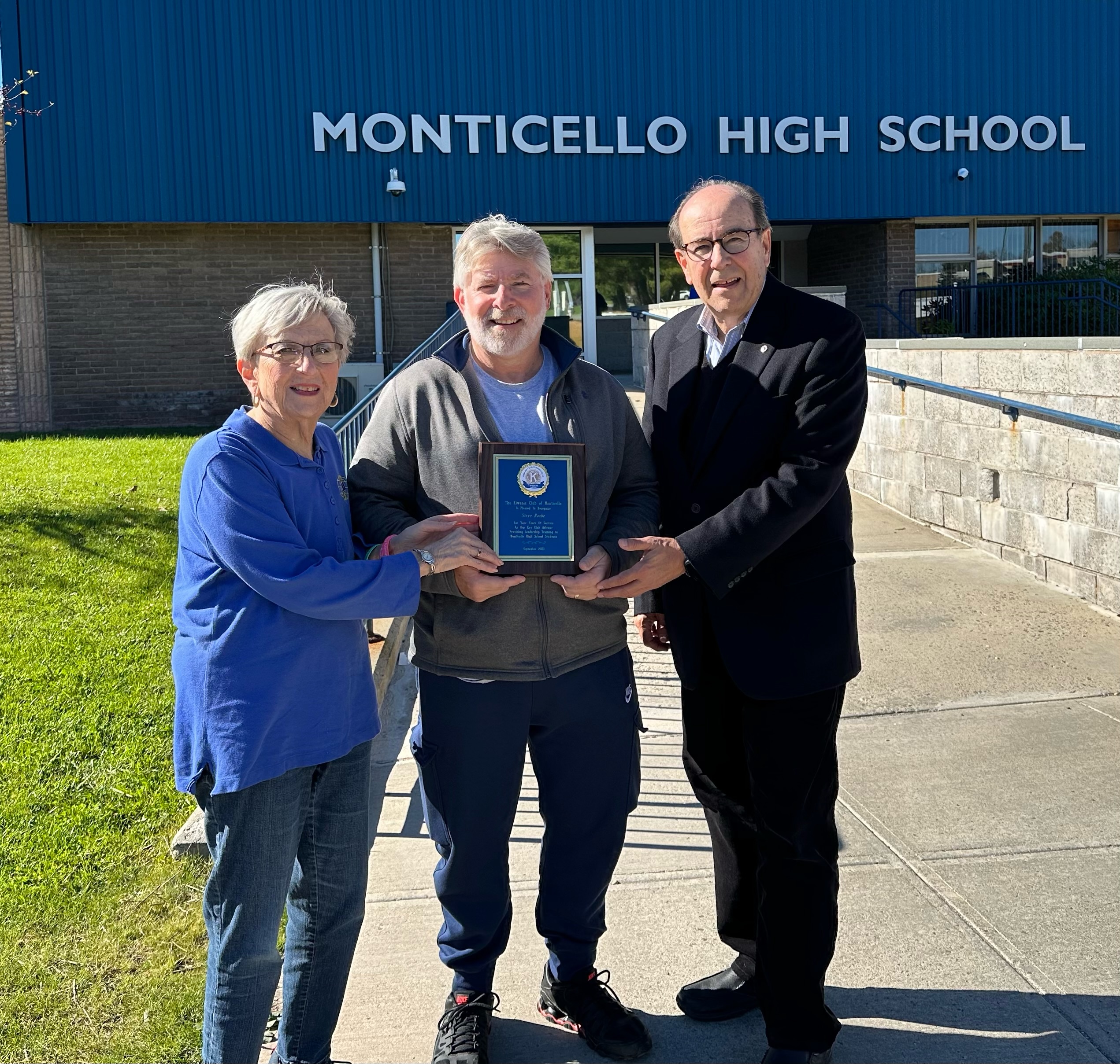 three people are standing in front of Monticello High School holding a plaque