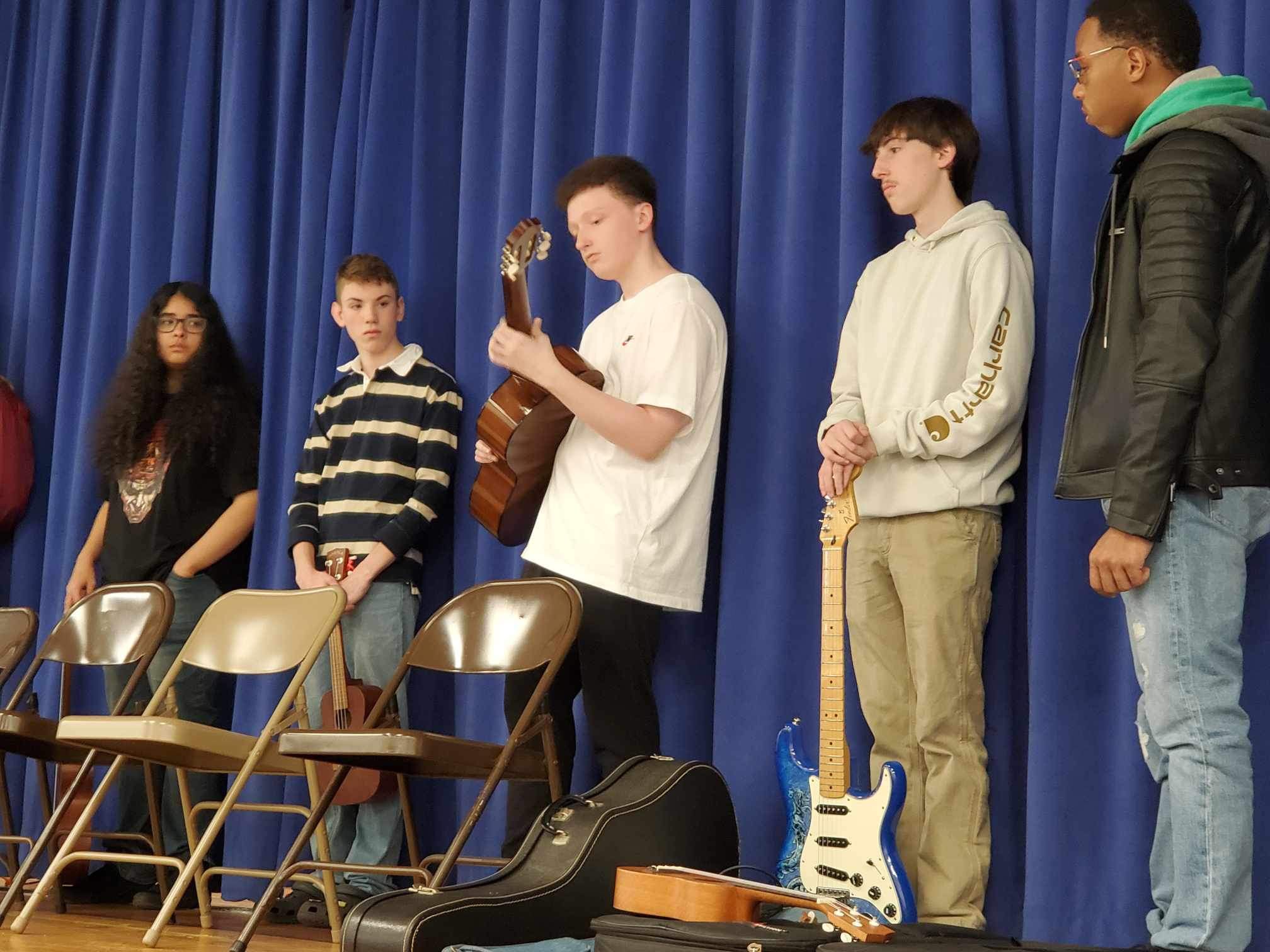 a student plays a guitar on stage as his peers look on. 