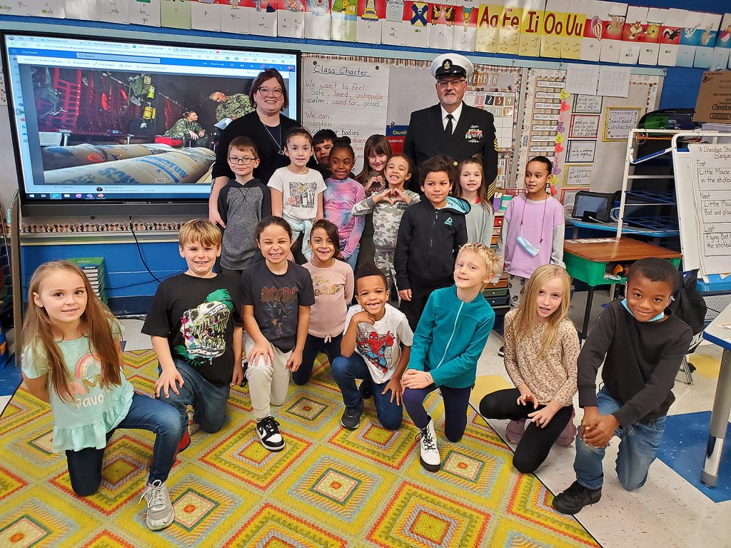 Man wearing a U.S. Navy uniform smiling for a group photo with an elementary teacher and her students.