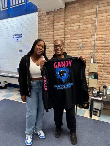 a staff member is showing off a shirt she was just given. A student is next to her and posing. 