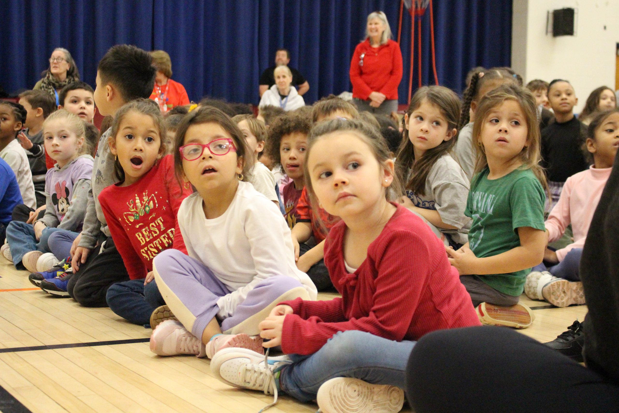 young students are seated on the floor of a gym. They are looking off camera and have a surprised look on their faces. 