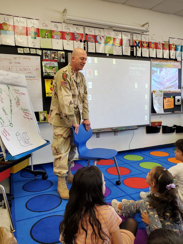 Man wearing a camouflage army uniform standing over a chair while speaking to elementary students.