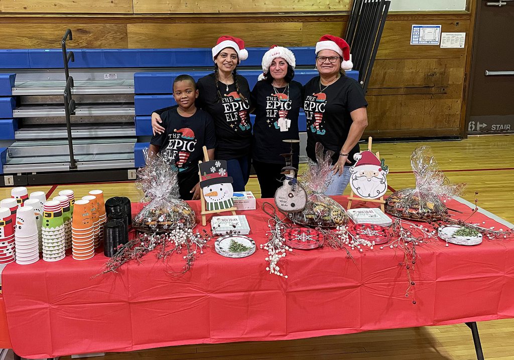 Three women and one student standing behind a table decorated with various holiday items.