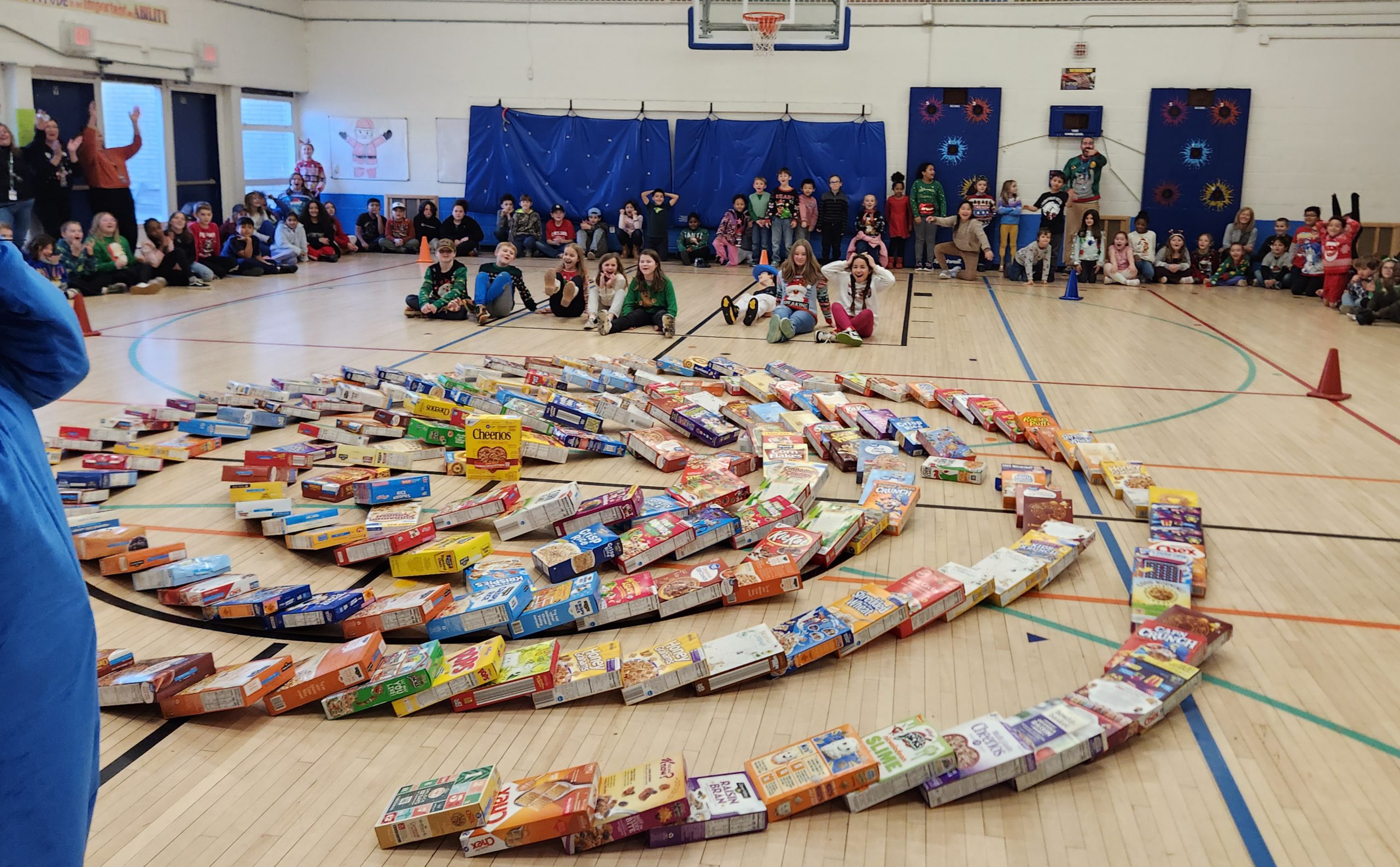 a group of students are seated on the floor of the gym watching cereal boxes fall. 