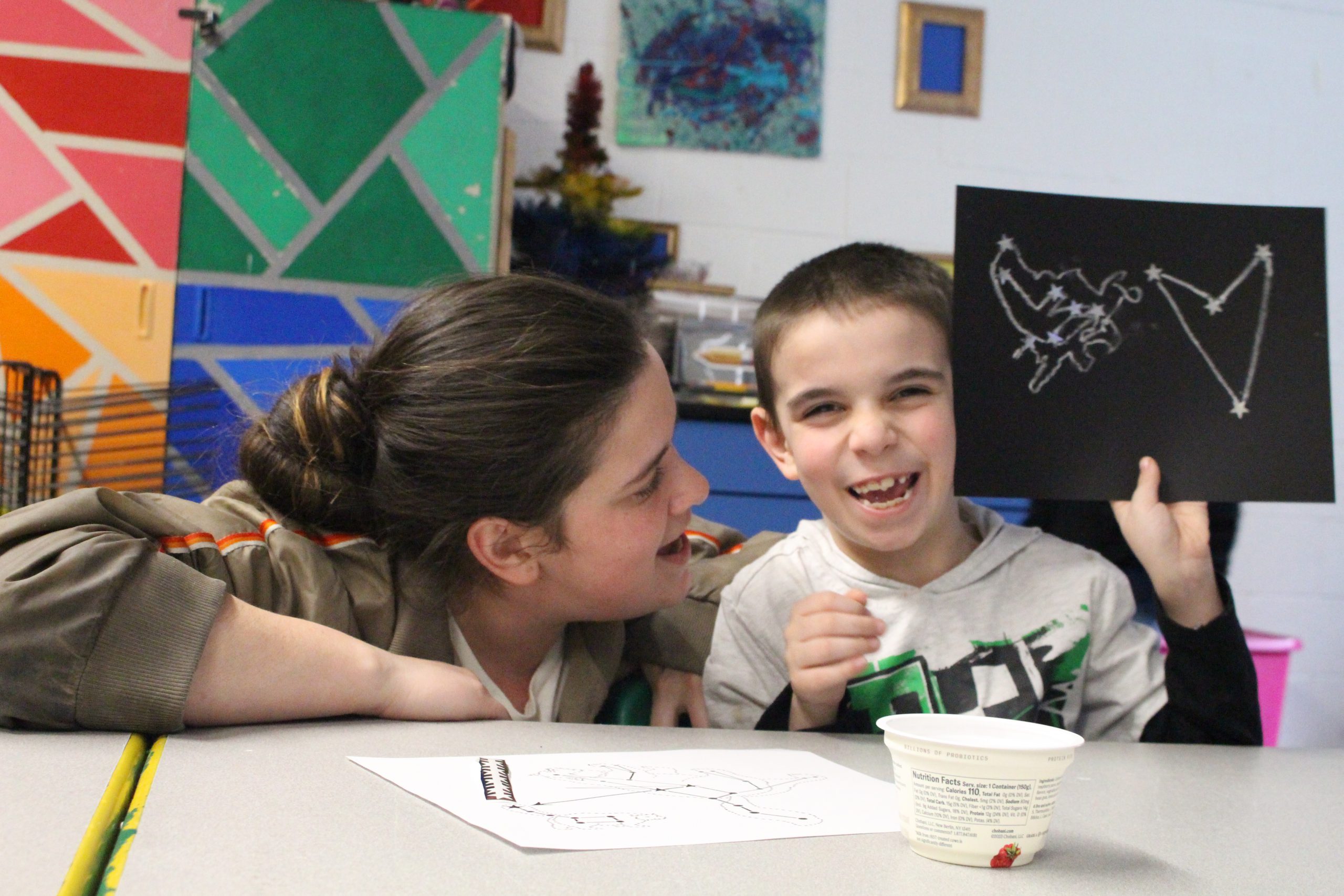 a child is holding up a drawing he made as his mother looks on. 