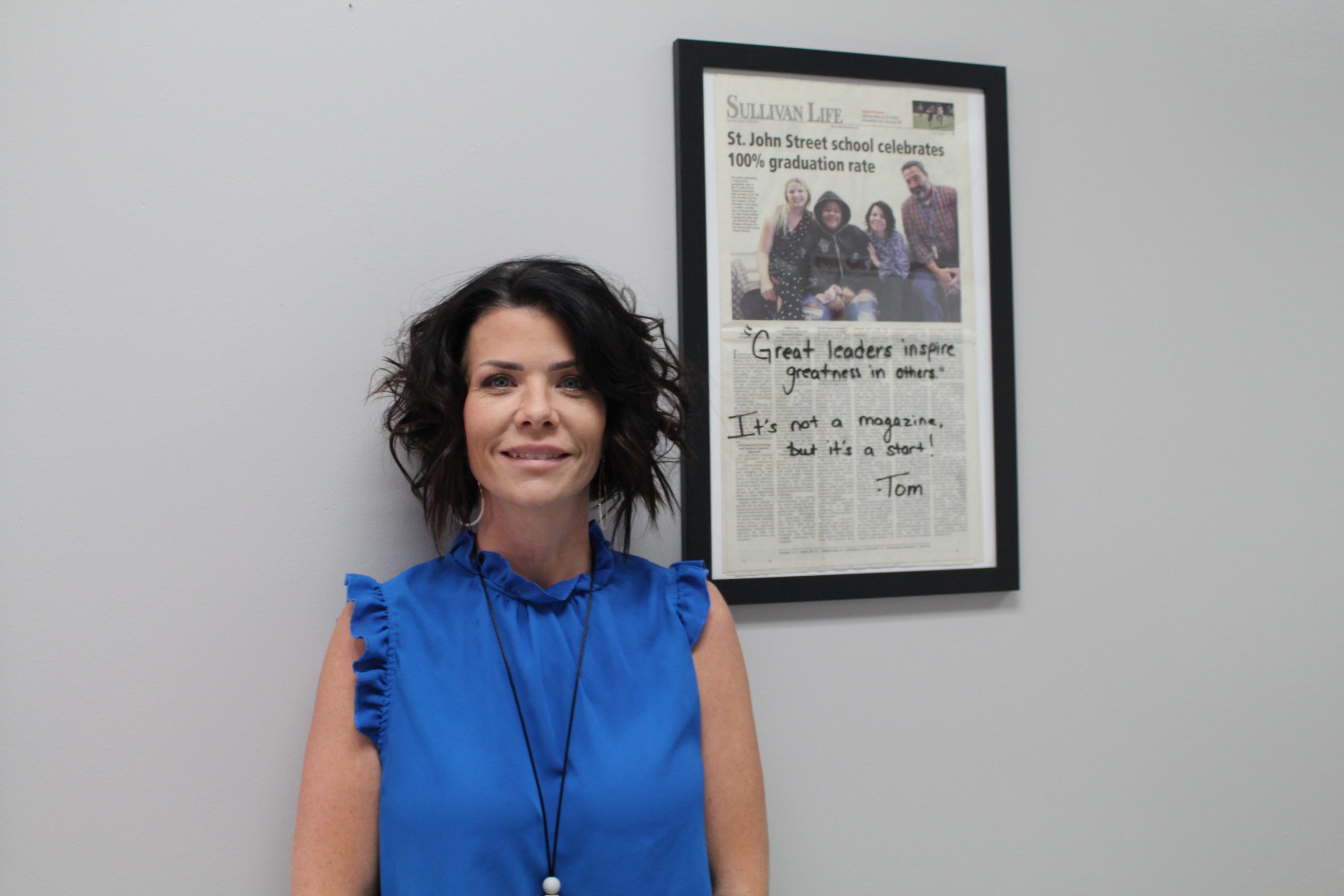 Jenn Gorr is posing by a framed article of her alternative program's 100% graduation rate. 