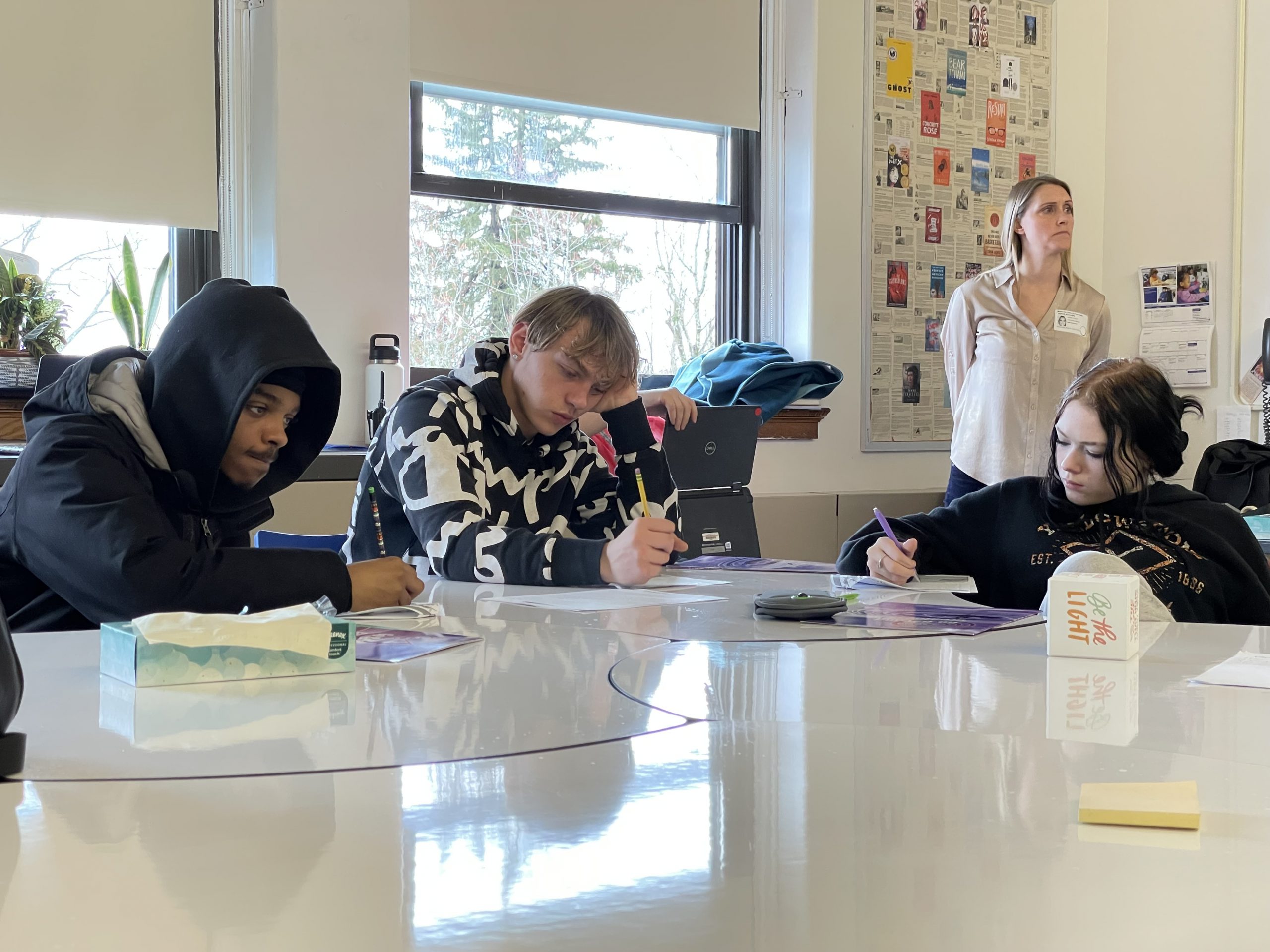 three students are seated at a desk, engaged in a writing activity. 