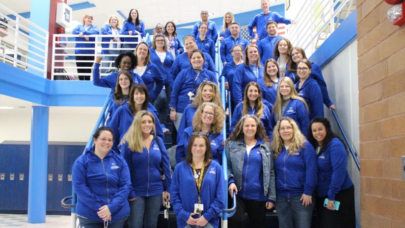 a large group of teachers is standing on the stairs and posing. They are all wearing matching blue sweatshirts.