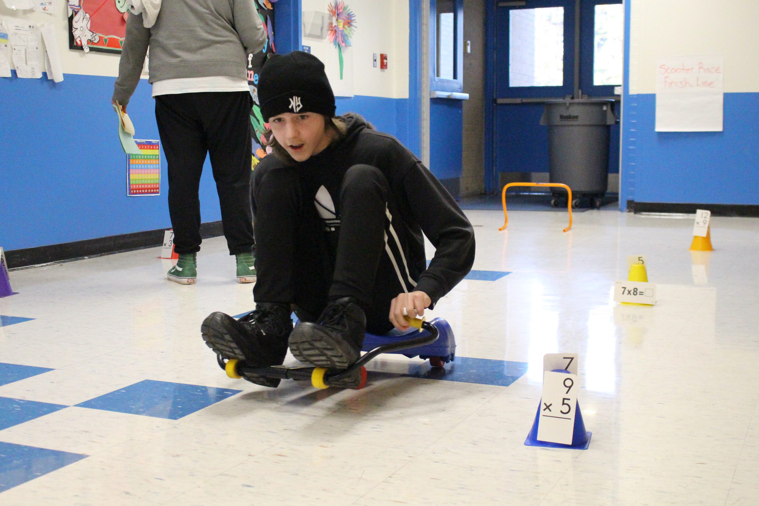 a student on a scooter is passing a math problem taped to a cone. 