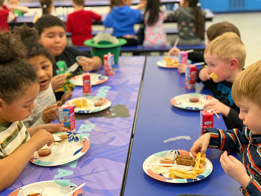 Elementary students eating waffles and sausage at a table.