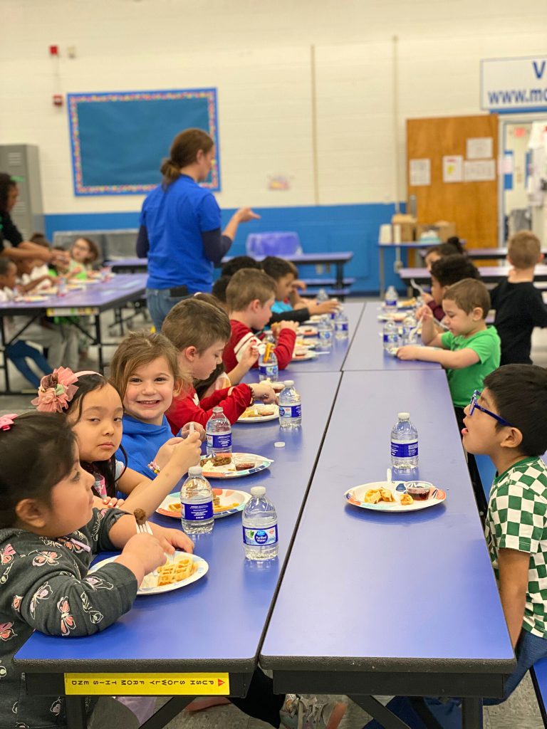 Elementary students sitting at a lunch table eating waffles and sausage.