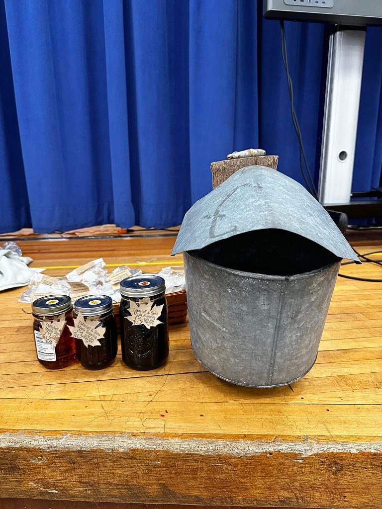 Three jars of maple syrup and a metal bucket displayed on the floor of the stage.