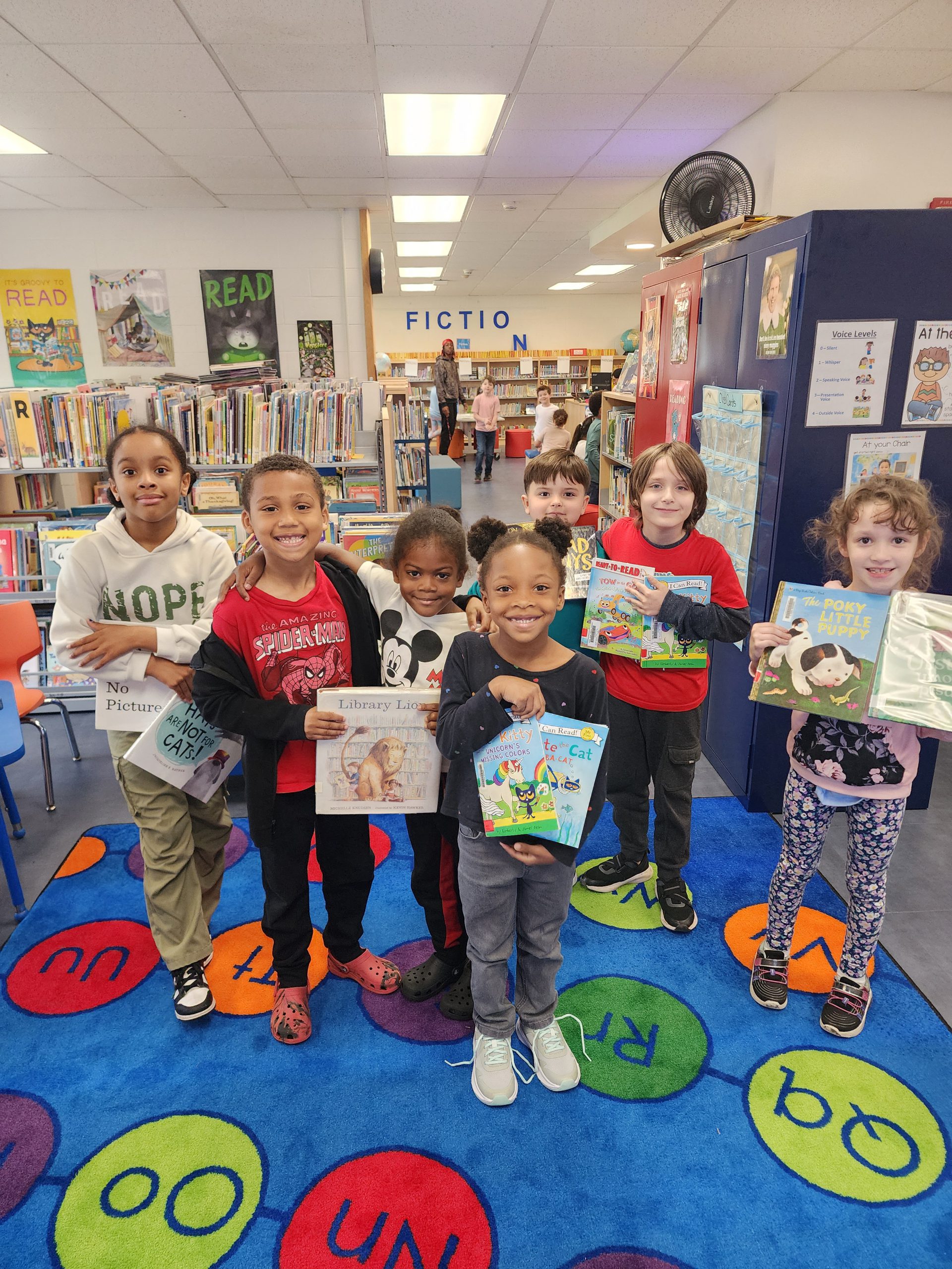 a group of young students are in a library, and holding up books smiling.