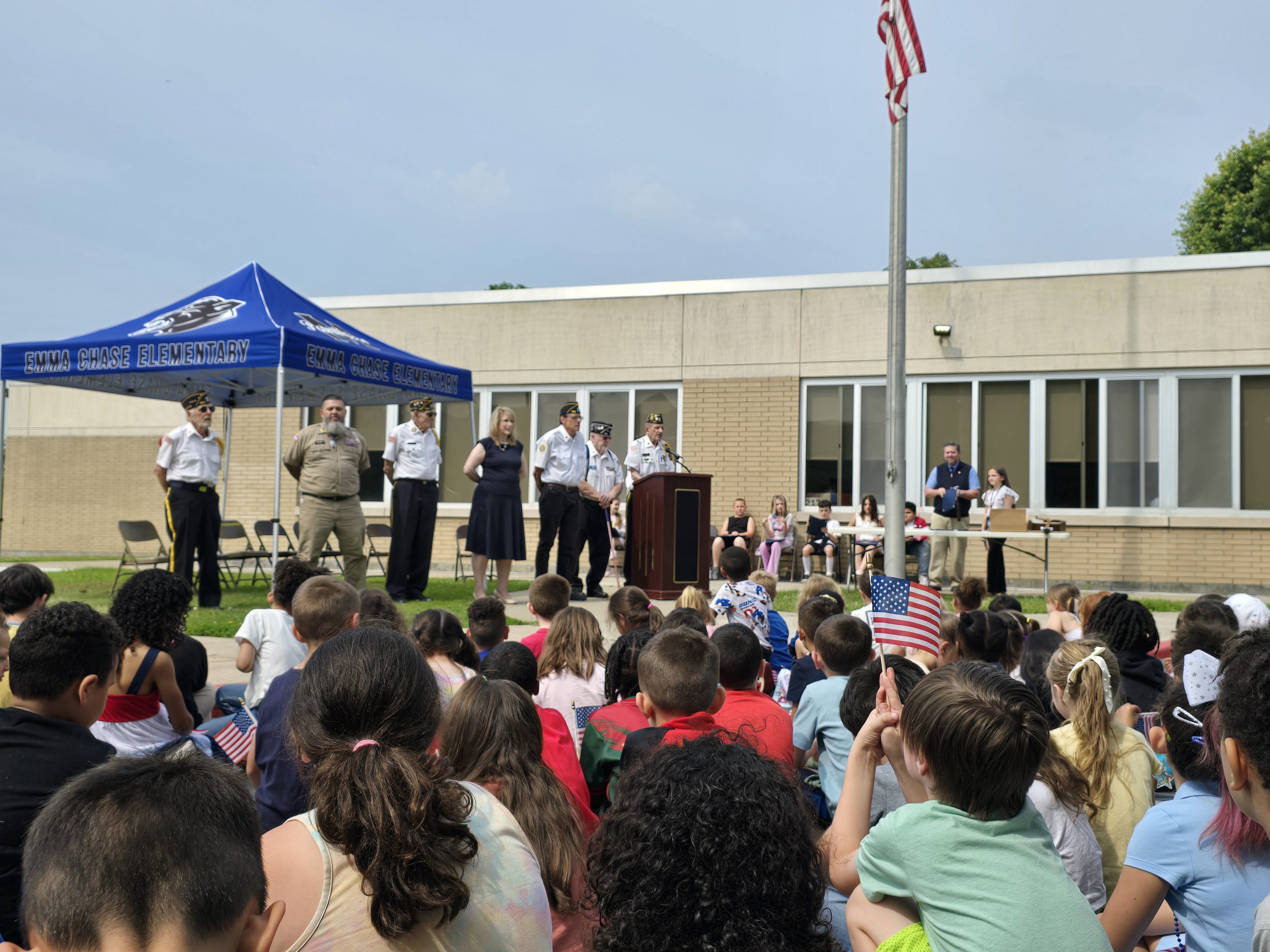 a large group of students are seated outdoors listening to veterans speaking