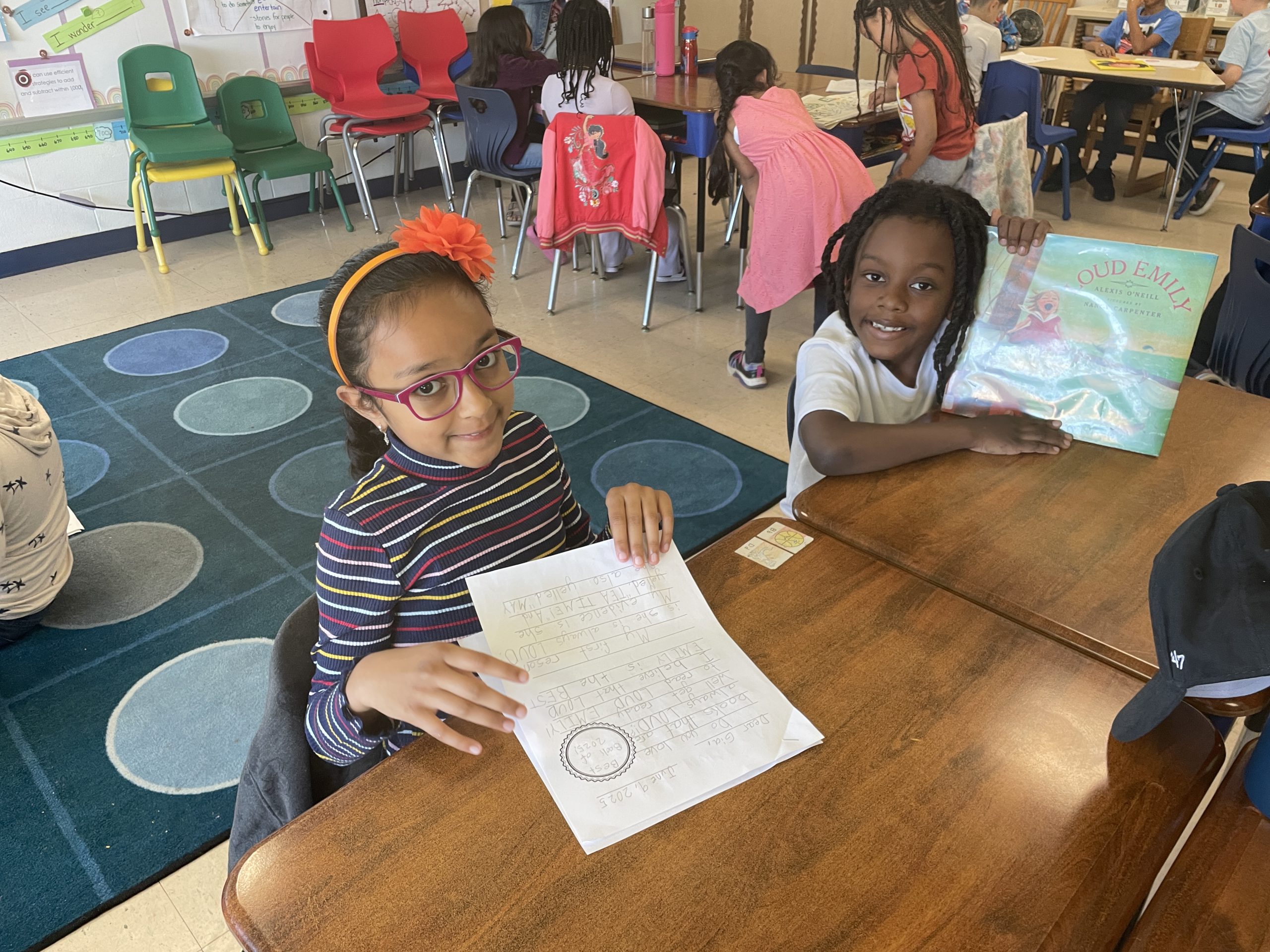 two students are seated at a desk smiling. 