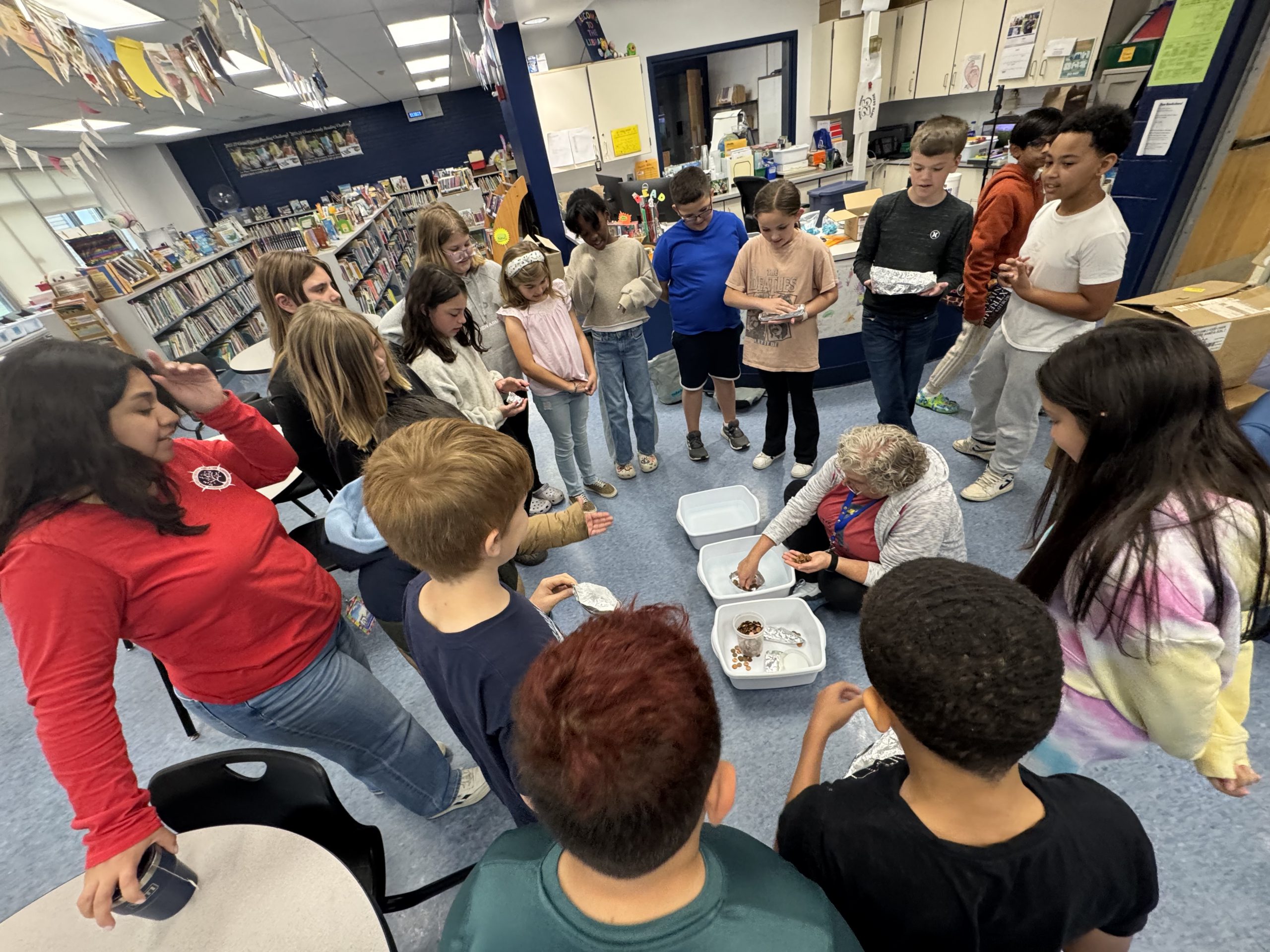 students surround Ms. Andersen as she places pennies in an alumnium foil boat in a tub of water 