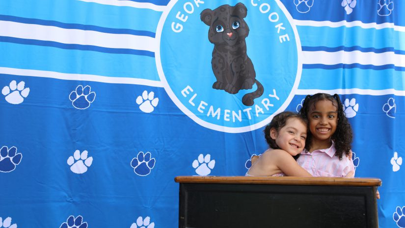 two young girls are smiling at a desk with a Cooke backdrop behind them