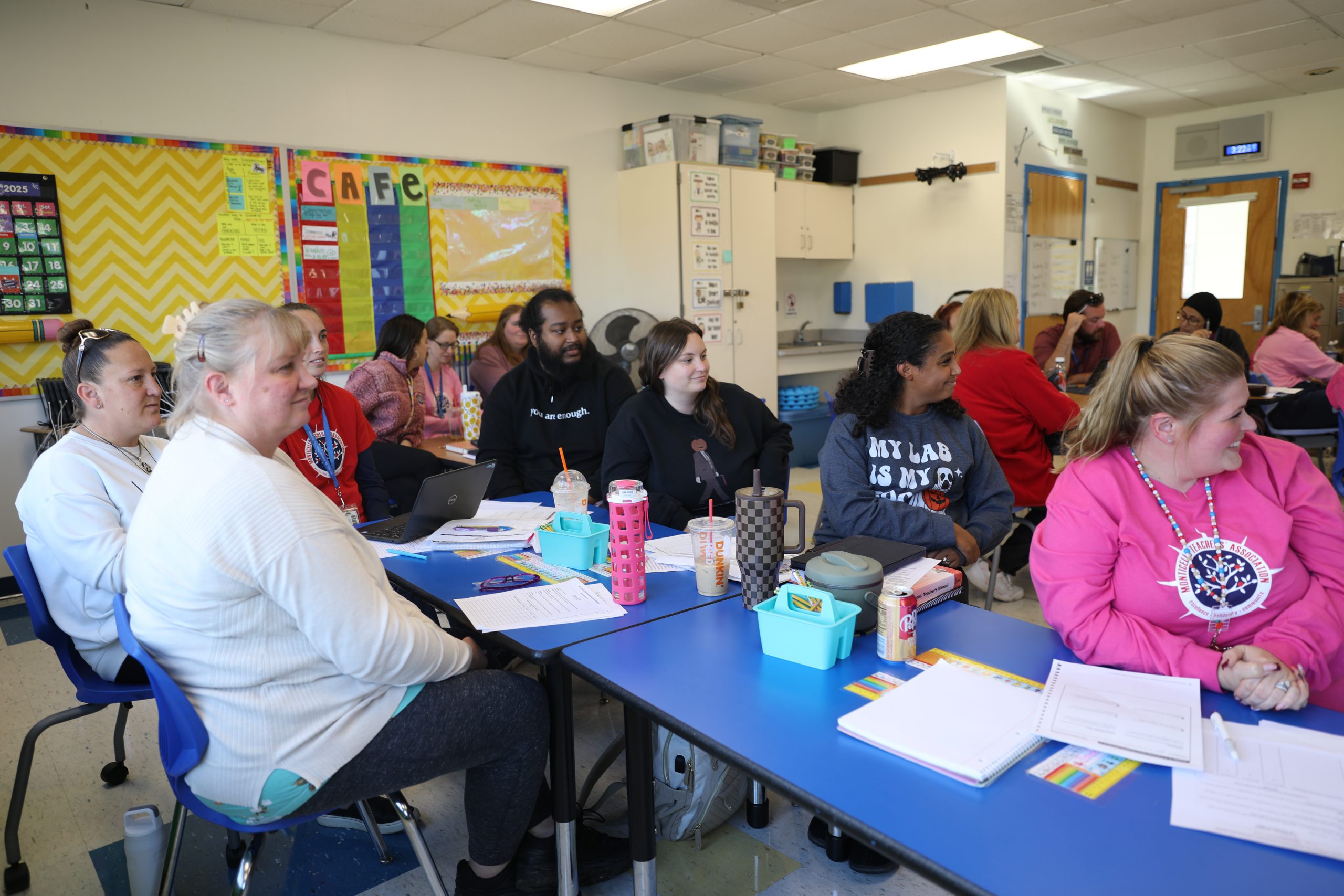 a group of teachers are seated at a table watching a presenter speak