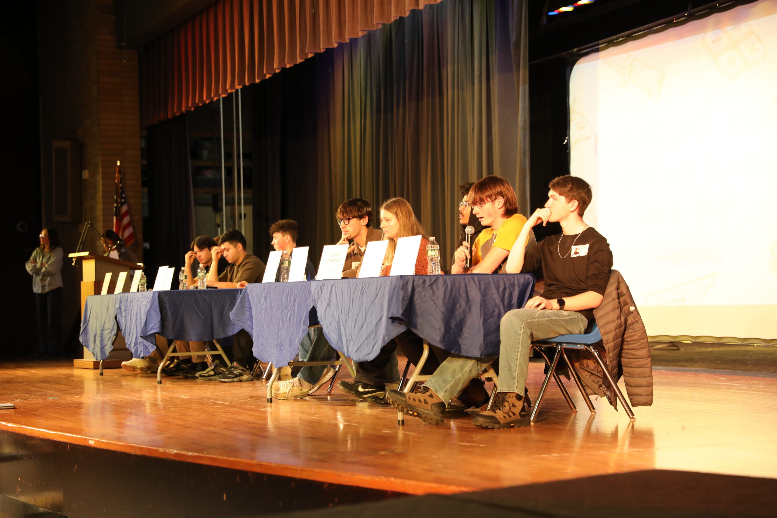 a group of alumni are seated on stage. one is speaking into the microphone