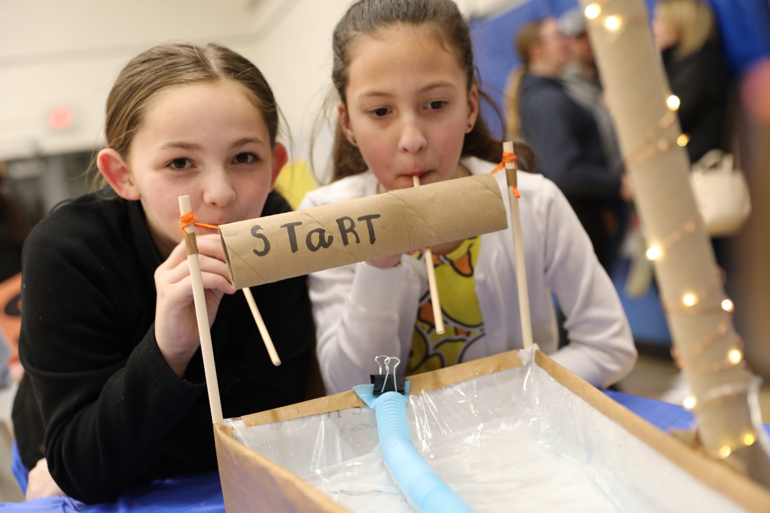 two students are playing a game where you blow through a straw to propel rubber ducks through water 