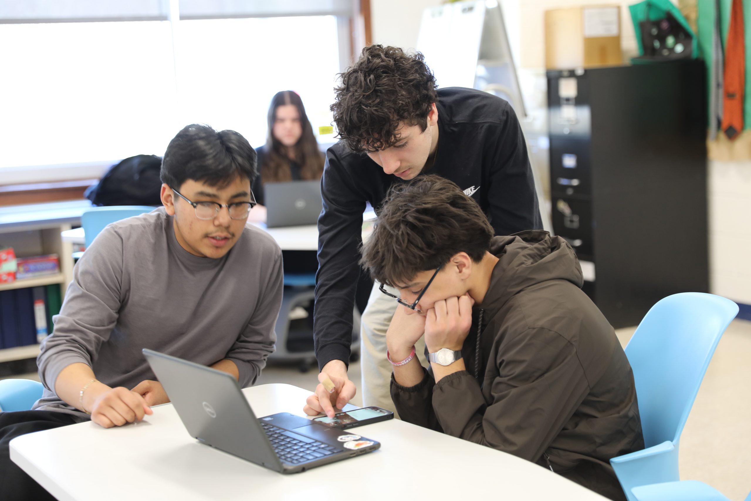 three students are gathered around a computer engaging in discussion