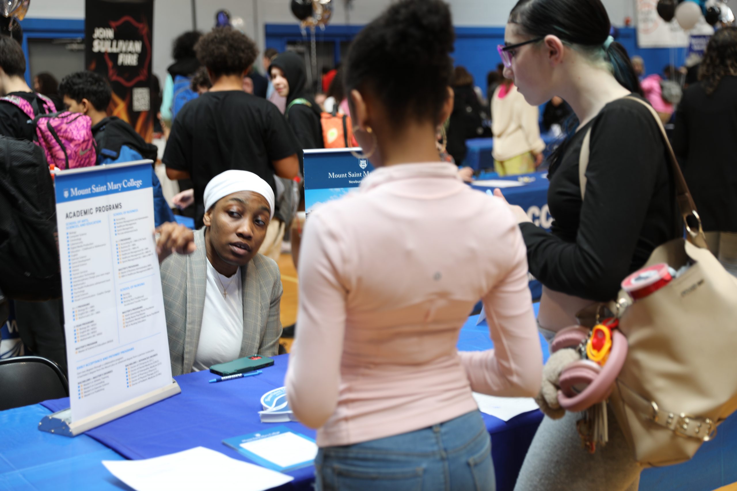 a vendor from a college points to an informational poster at her booth. Two students are looking on. 