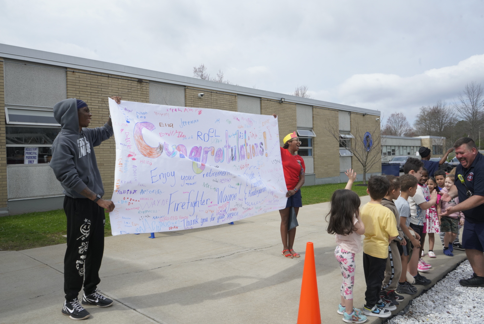 Two staff members hold up a large sign that reads "congratulations" while firefighter Wayne greets students lined up on the sidewalk