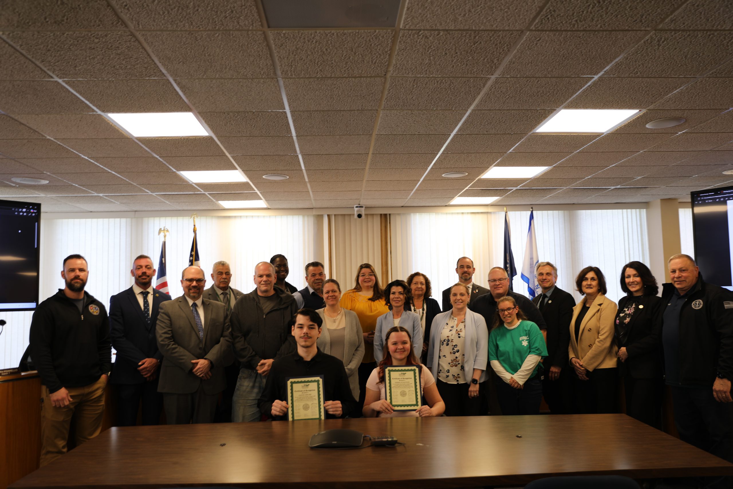 Gavin and Emma are seated at a table, holding up certificates and surrounded by various legislators