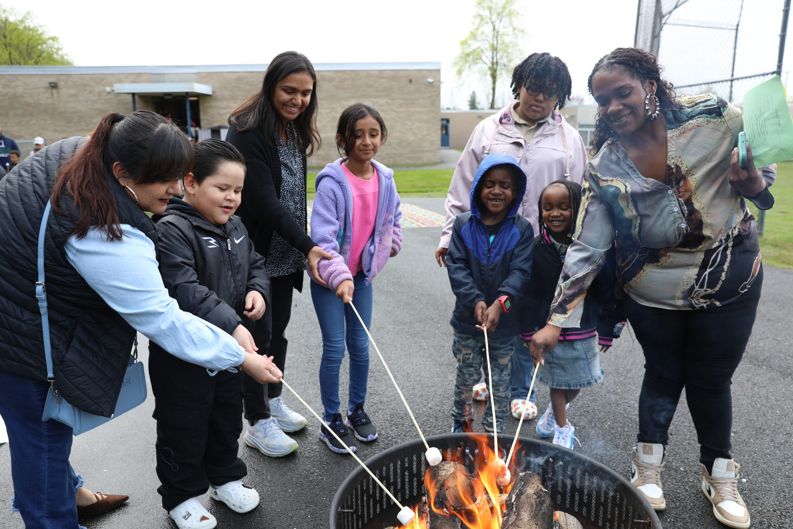 a group of families are gathered around an outdoor bonfire roasting marshmellows