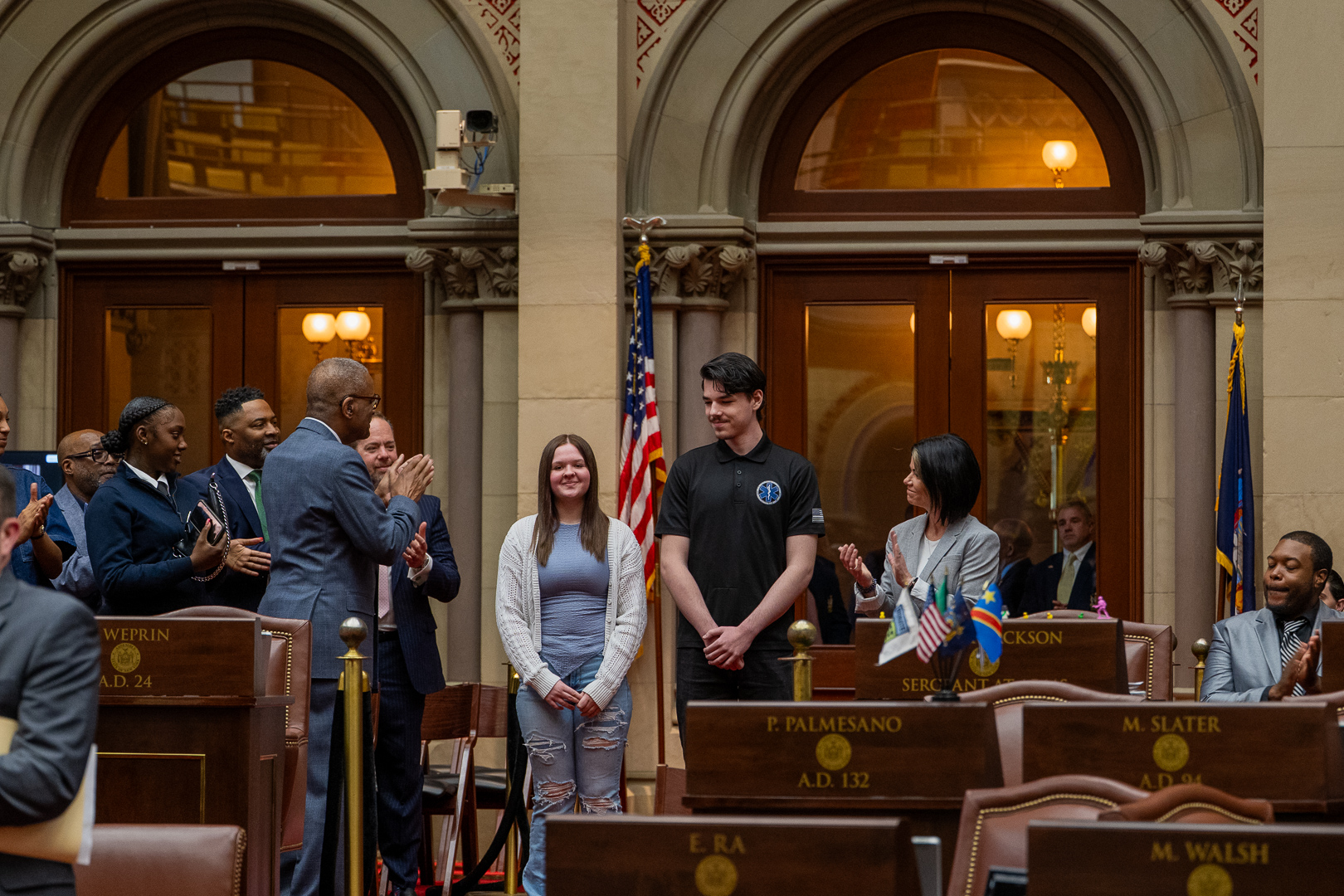 Gavin and Emma are standing in the center of a group of Assembly members who are breaking out in applause 