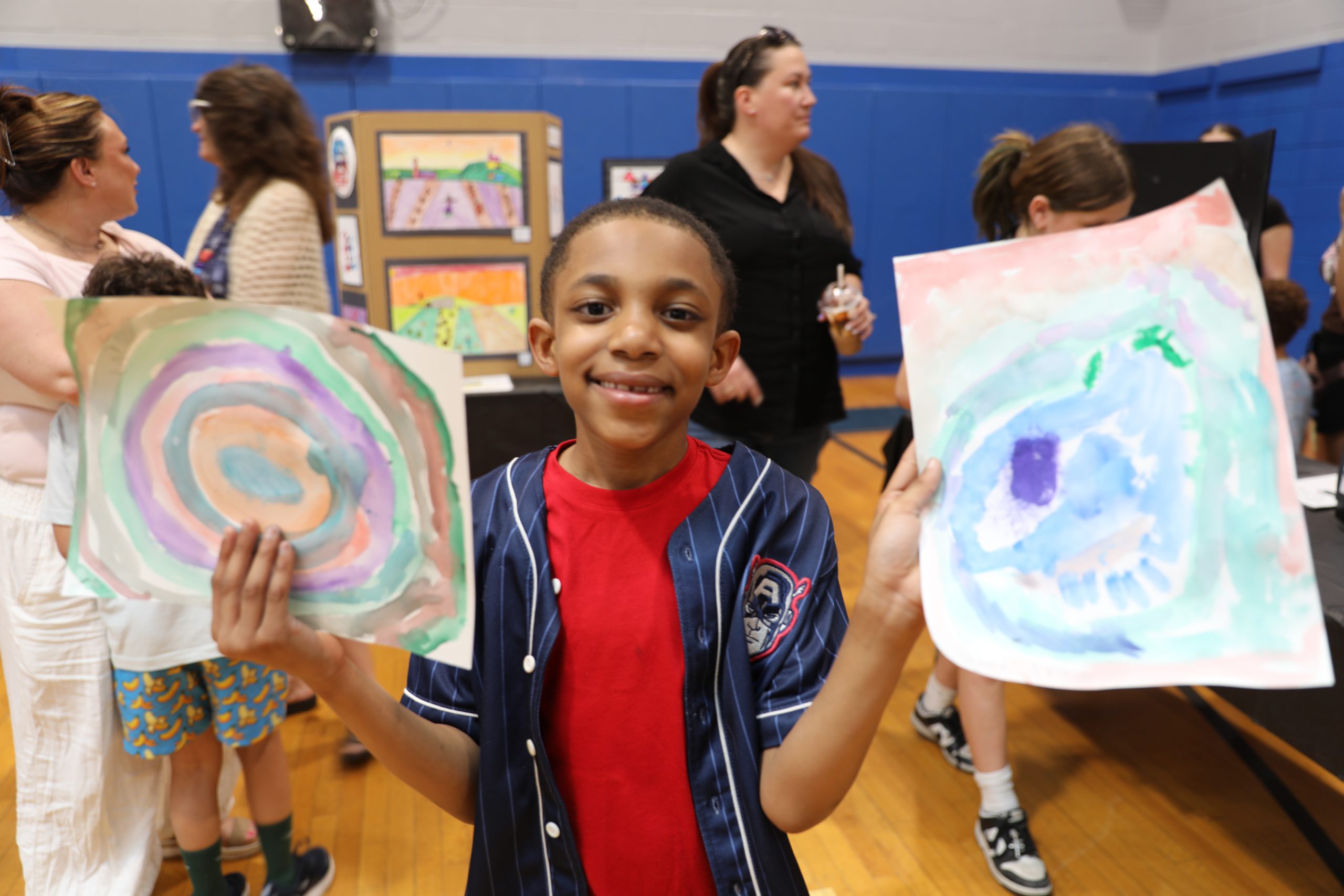 a student holds up two crayon drawings, one in each hand, and smiles for the camera.