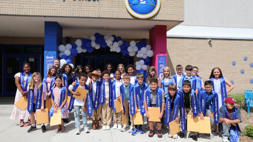 the fifth grade students, wearing blue stolls, are all gathered in a group posing for the camera under a celebratory balloon arch