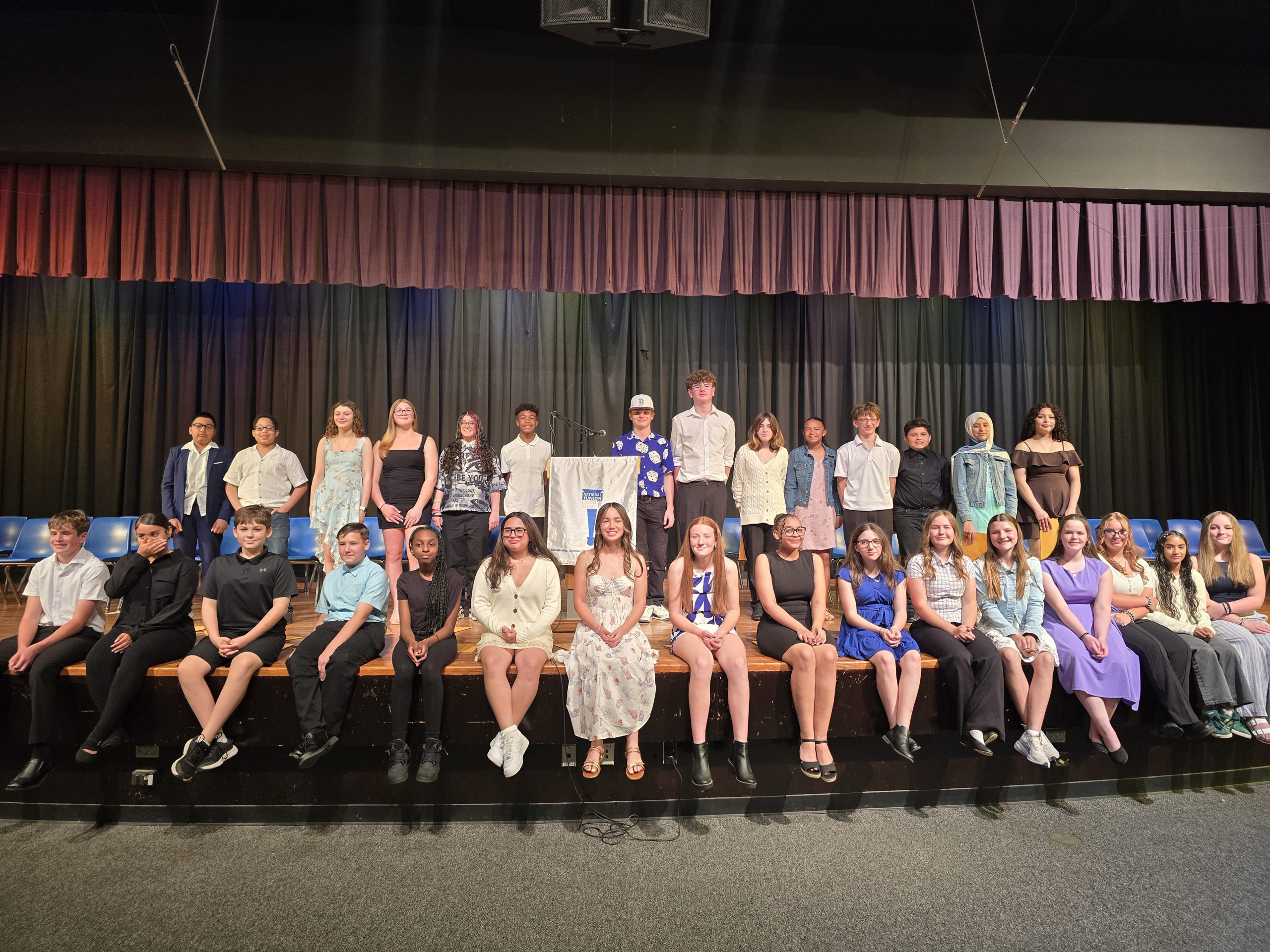 the students who were inducted into the National Junior Honor Society are seated on stage, posing for the camera. 