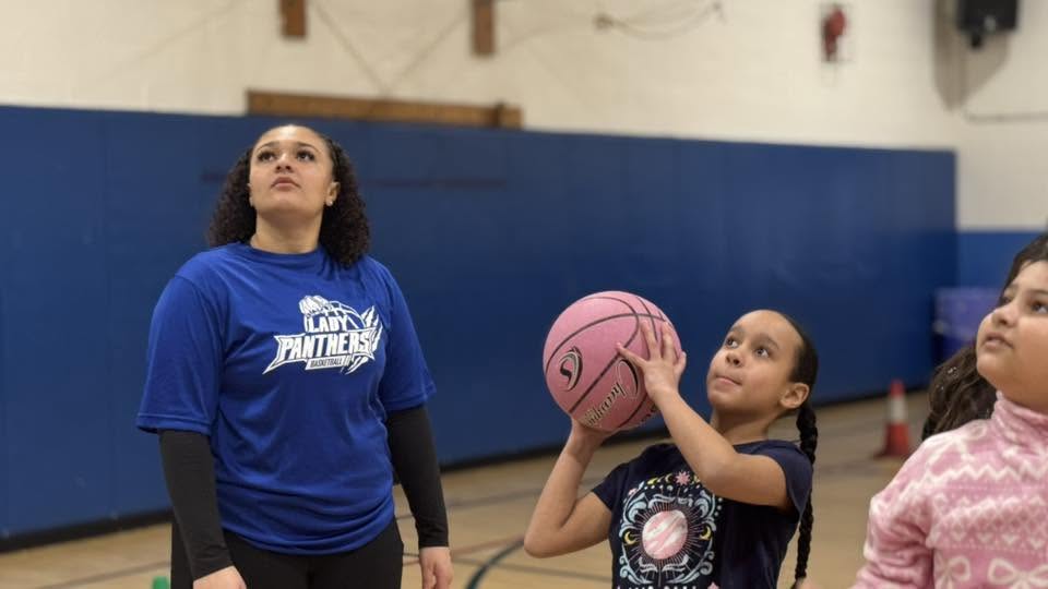 an elementary student is poised to throw a basketball as a senior athlete looks on. 
