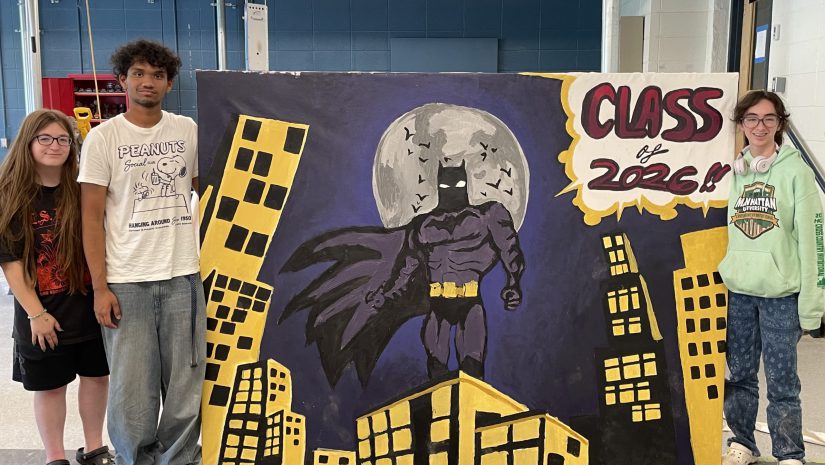 three students are standing next to the mural they painted. The mural features batman standing on top of a building and the words class of 2026