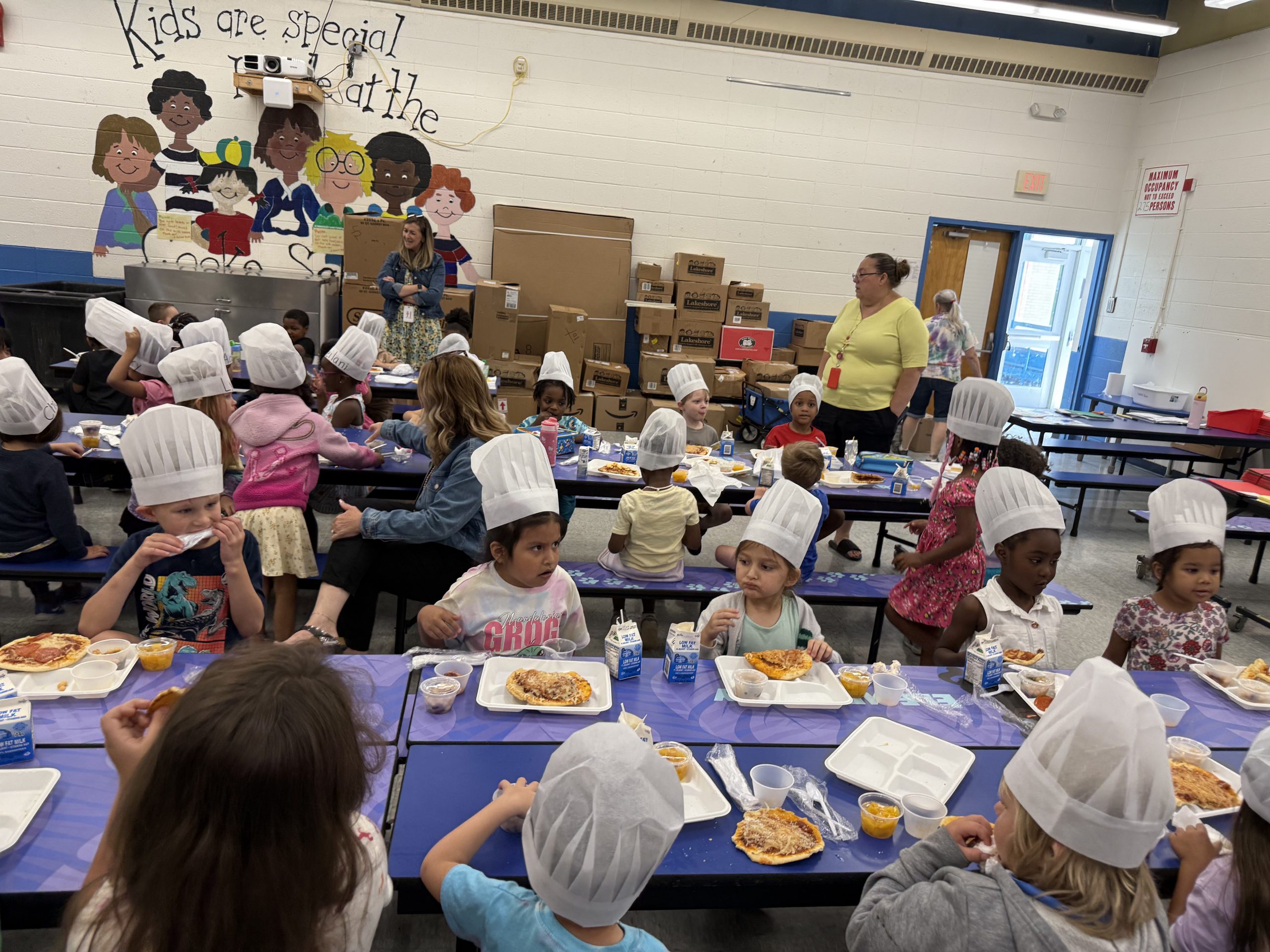 a group of young students eating pizza are seated in a cafeteria. All are wearing chef hats