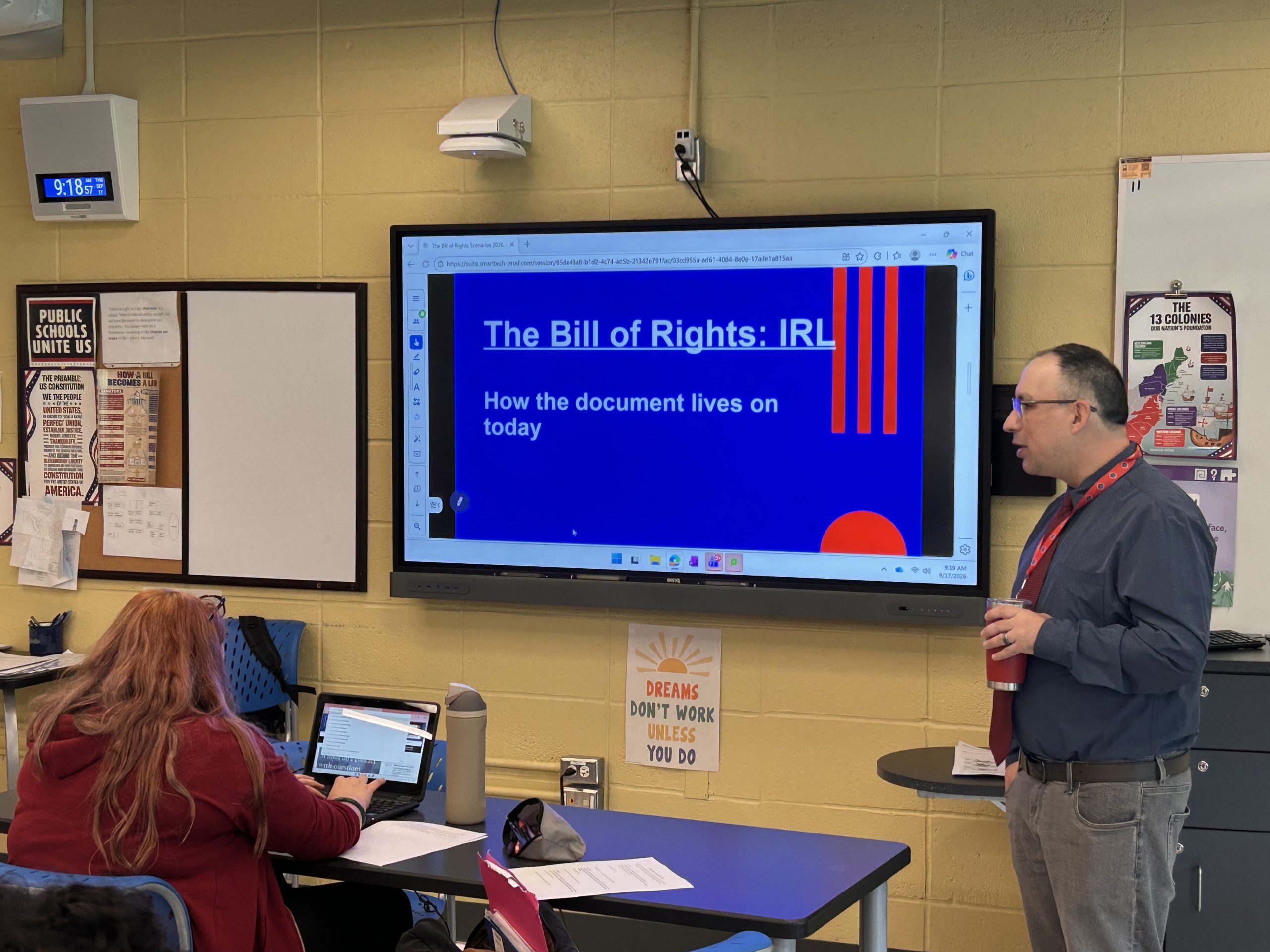 a teacher addresses students in class. On the whiteboard is a slide that says The Bill of Rights
