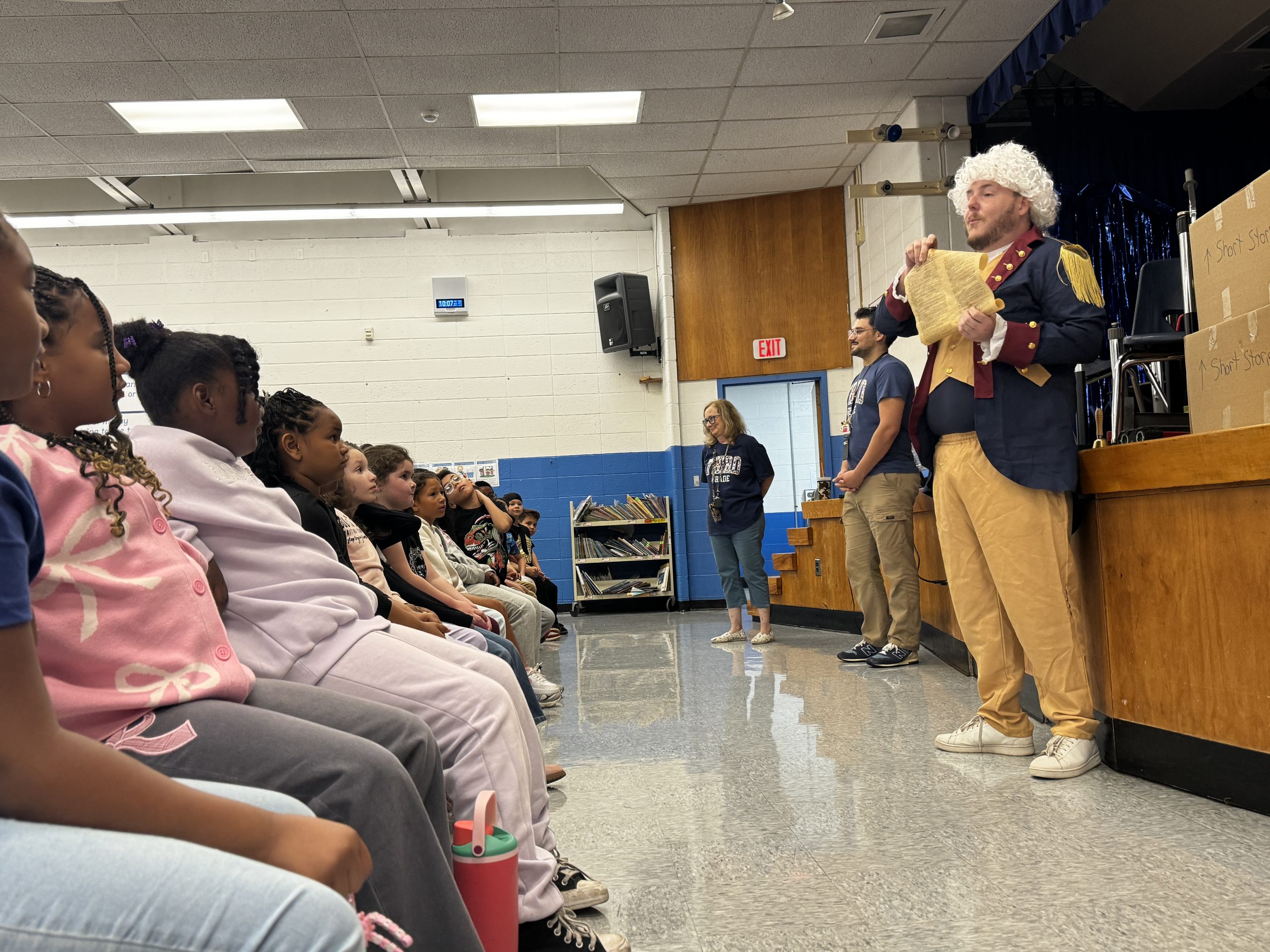 A teacher dressed as George Washington is holding a replica of the constituiton and addressing a room of students