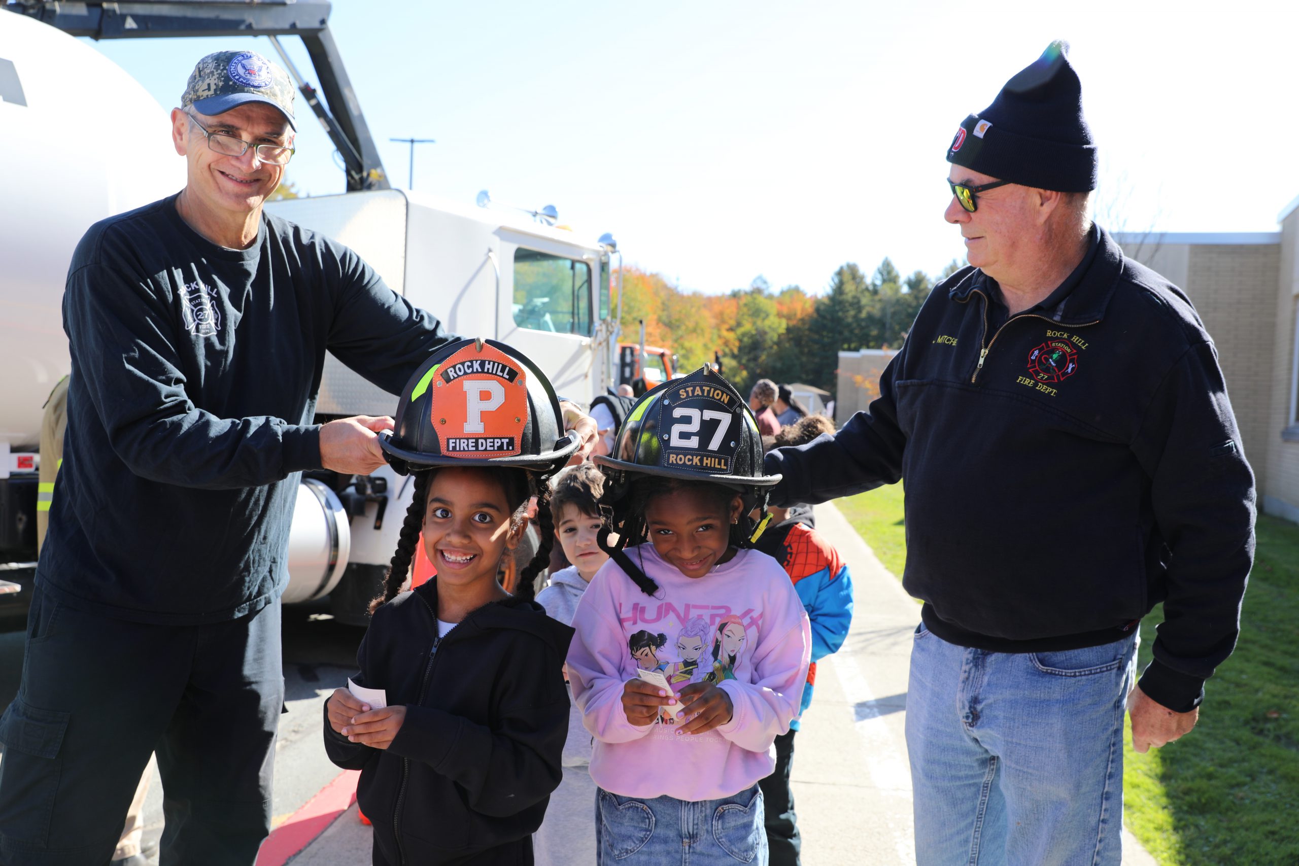 two firefighters are helping students try on fire hats during touch a truck day